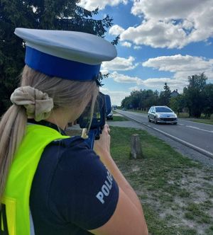 Policjantka trzyma urządzenie do pomiaru prędkości skierowane na drogę, którą porusza się pojazd